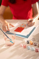 Brush resting in a glass of water beside numbered paint pots, with the Fotomatic canvas being painted in the background