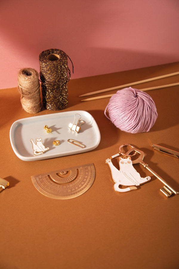 White cat enamel pin on a ceramic tray next to the matching "Don't give a sh*t" cat keychain, with yarn and knitting needles