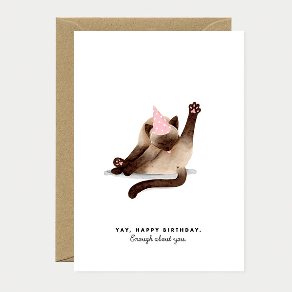 Illustrated birthday greeting card of a Siamese cat wearing a pink polka dot party hat, grooming itself with one leg raised, text reading "Yay, happy birthday. Enough about you."