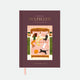 A5 Hardcover notebook with a hand-painted illustration of a woman in bed with four cats and a dog on a burgundy background by All the Ways to Say.