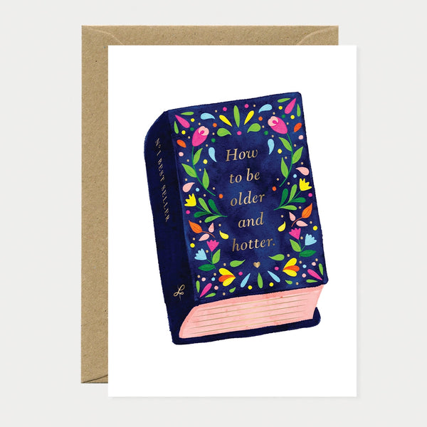 Illustrated birthday greeting card designed as a navy book cover surrounded by colorful flowers, gold foil text reading "How to be older and hotter" and "N°1 Bestseller" along the spine