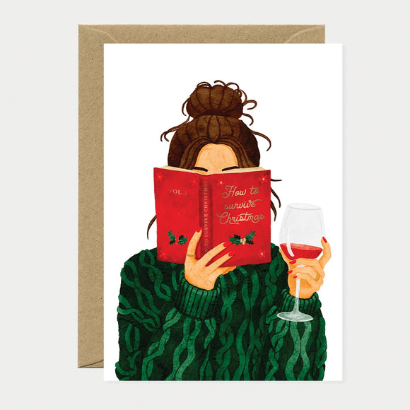 Illustrated Christmas card of a woman in a green sweater reading a red book titled "How to Survive Christmas" while holding a glass of red wine