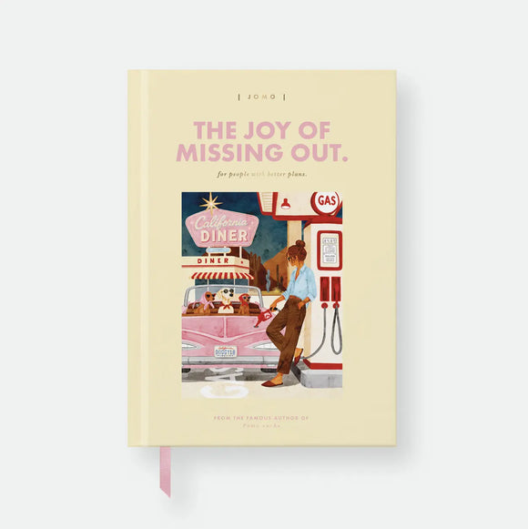 JOMO hardcover A5 notebook by All The Ways To Say, pale yellow cover styled like a book jacket with the title "The Joy Of Missing Out, for people with better plans," a fake author credit, and an illustration of a woman pumping gas at a retro gas station with three dogs in a pink convertible