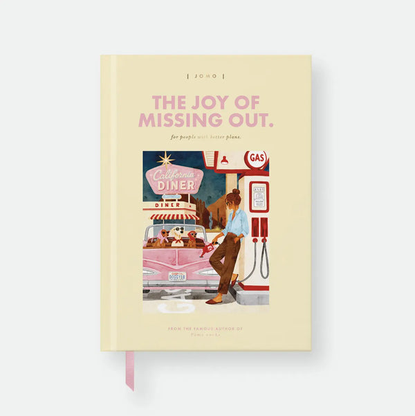 JOMO hardcover A5 notebook by All The Ways To Say, pale yellow cover styled like a book jacket with the title "The Joy Of Missing Out, for people with better plans," a fake author credit, and an illustration of a woman pumping gas at a retro gas station with three dogs in a pink convertible