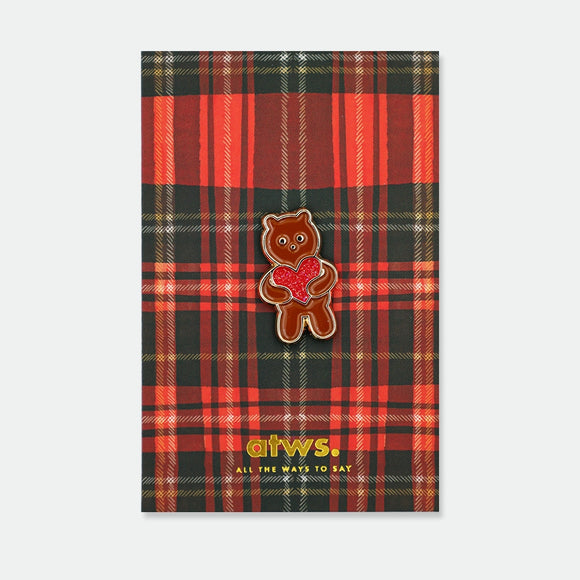 Enamel pin of a brown bear standing upright, holding a red glitter heart with both paws, gold outline, on an illustrated tartan backing card