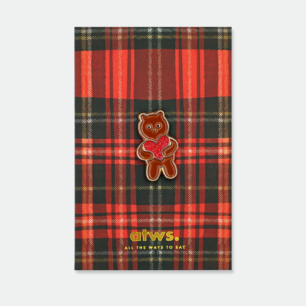 Enamel pin of a brown bear standing upright, holding a red glitter heart with both paws, gold outline, on an illustrated tartan backing card