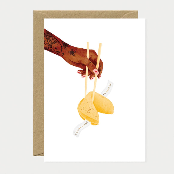 Illustrated greeting card of a tattooed hand holding chopsticks over an open fortune cookie, gold foil text on the fortune reading "You're lucky to have me" and "destiny" written on the chopstick