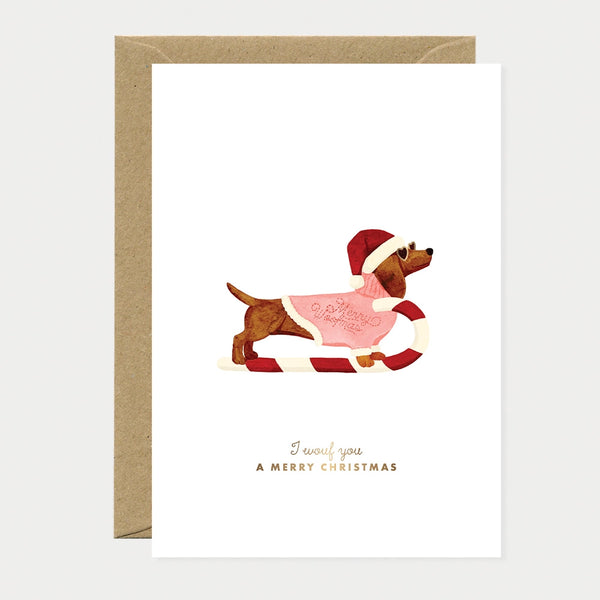 Illustrated Christmas card of a dachshund shaped like a candy cane sled, wearing a Santa hat, heart-shaped sunglasses, and a pink sweater reading "Merry Woofmas," gold foil text reading "I wouf you a Merry Christmas"