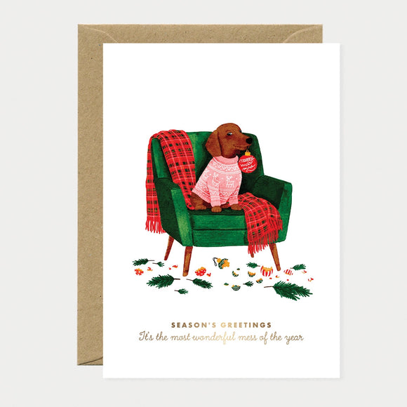 Illustrated Christmas card of a dachshund wearing a pink reindeer-print sweater, sitting in a green armchair with a red plaid throw and an ornament tag reading "Merry Woof-mas," surrounded by broken ornaments, gold foil text reading "Season's Greetings, It's the most wonderful mess of the year"