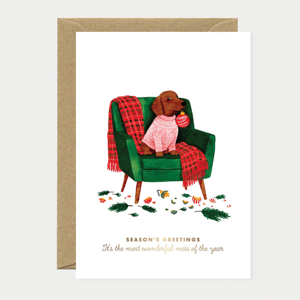 Illustrated Christmas card of a dachshund wearing a pink reindeer-print sweater, sitting in a green armchair with a red plaid throw and an ornament tag reading "Merry Woof-mas," surrounded by broken ornaments, gold foil text reading "Season's Greetings, It's the most wonderful mess of the year"