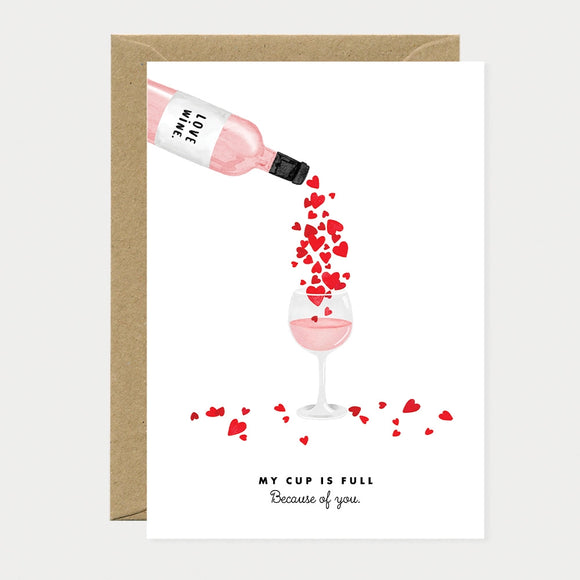 Illustrated greeting card of a bottle labeled "Love Wine" pouring red hearts into a wine glass, with hearts scattered around the base, text reading "My cup is full, Because of you"