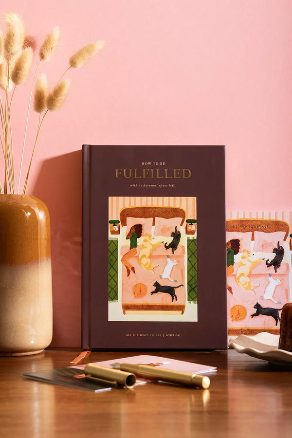 How to be fulfilled Hardcover Notebook A5 - Front view on a wooden desk with a dried flower vase.