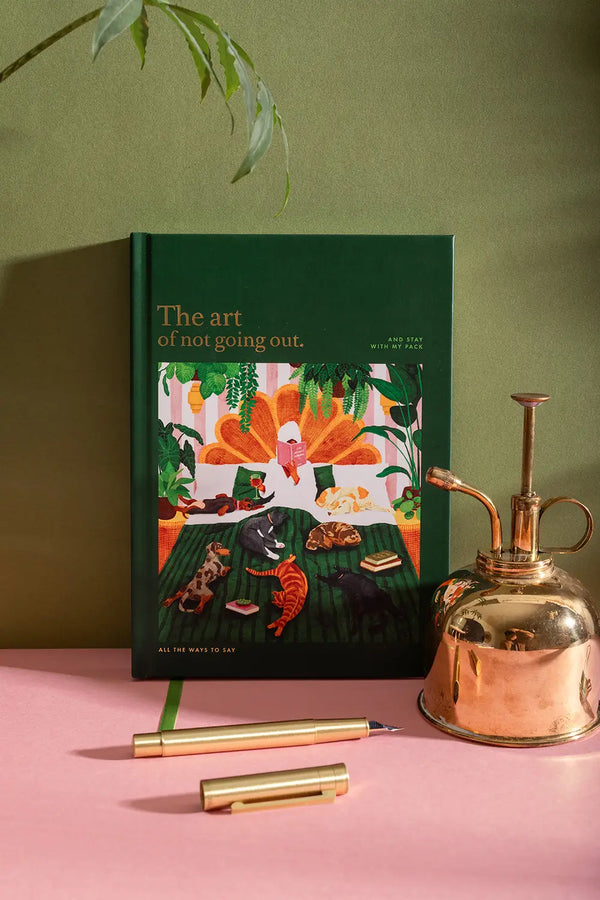 The art of not going out Hardcover Notebook A5 - Front view on a pink desk with a gold water sprayer.