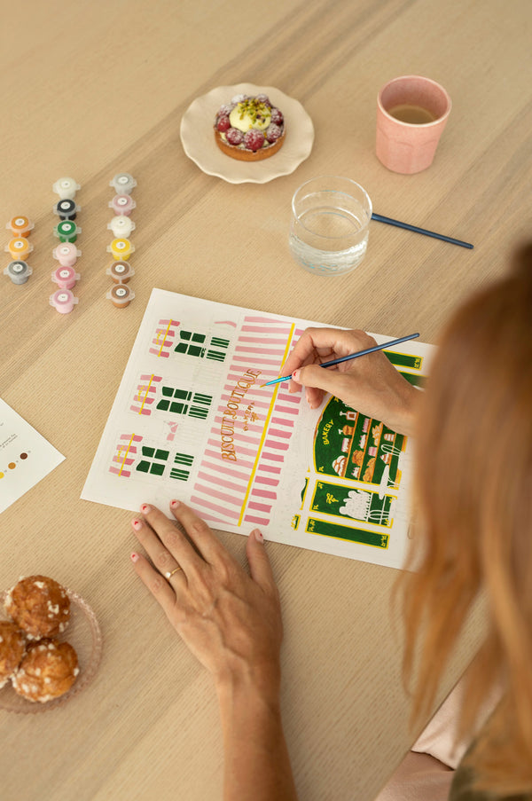 Hand painting the Pink Bakery paint by numbers canvas in pink, green and gold, with paint pots and a raspberry tart