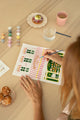 Hand painting the Pink Bakery paint by numbers canvas in pink, green and gold, with paint pots and a raspberry tart