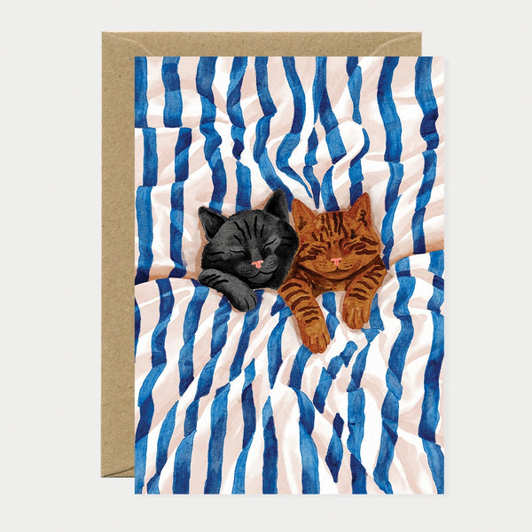 Illustrated greeting card of a black cat and a tabby cat sleeping together on blue and white striped sheets