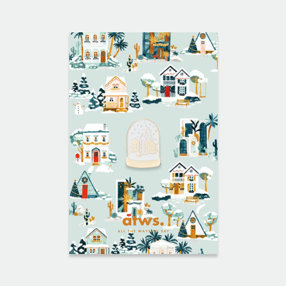 Chic Snowball enamel pin by All the Ways to Say featuring golden snow globe design with small winter village on illustrated pastel background with houses and snow.
