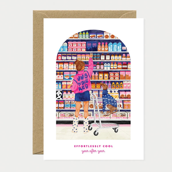 Illustrated birthday greeting card of a woman in a pink jacket reaching for a shelf in a candy and grocery aisle, with a dachshund sitting in the shopping cart below, text reading "Effortlessly cool, year after year"
