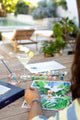 Painting the Swing paint by numbers kit on an outdoor table by the pool, with a cocktail and a glass of water