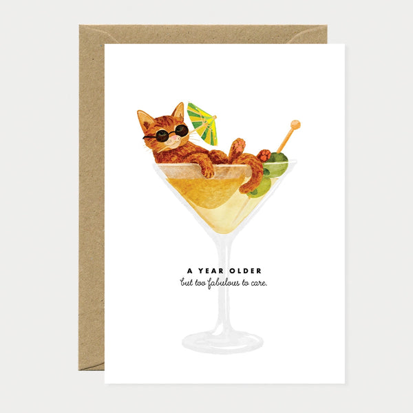 Illustrated birthday greeting card of a ginger cat wearing sunglasses, lounging inside a martini glass with a cocktail umbrella and olives, text reading "A year older, but too fabulous to care"