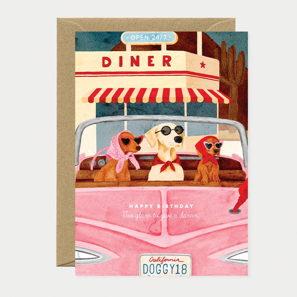 Illustrated birthday greeting card of three dogs wearing sunglasses and headscarves riding in a pink convertible outside a roadside diner, license plate reading "California DOGGY18," text reading "Happy birthday, Too glam to give a damn"