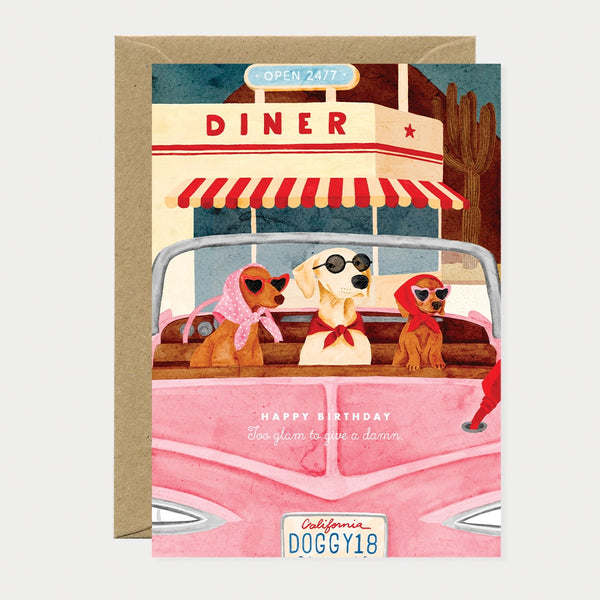 Illustrated birthday greeting card of three dogs wearing sunglasses and headscarves riding in a pink convertible outside a roadside diner, license plate reading "California DOGGY18," text reading "Happy birthday, Too glam to give a damn"