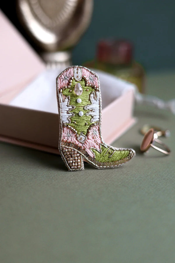 Bohemian style cowboy boot jewelry piece styled with gold accessories and a decorative hand sculpture in the background.