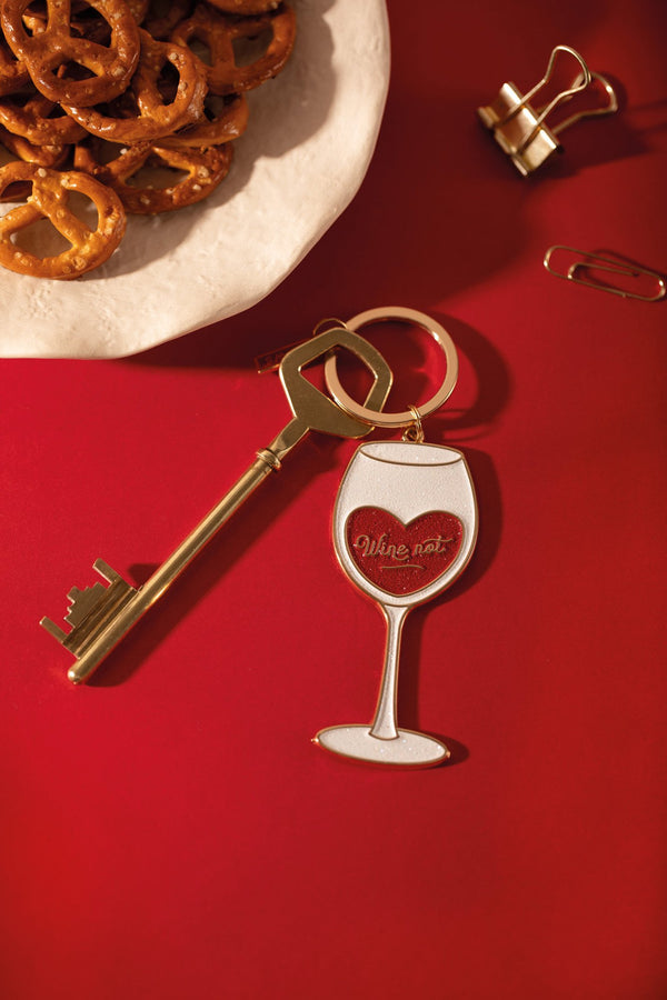"Wine not" enamel keychain shaped like a glittery wine glass with a red heart, next to a gold key and pretzels on a red background
