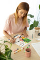 Woman painting the Dog Sitter paint by numbers kit at a wooden table with plants, paint pots and a cookie