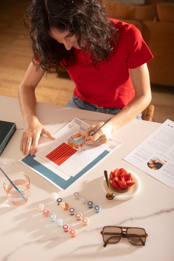 Woman painting the Fotomatic paint by numbers kit on a marble table with paint pots, a strawberry tart and sunglasses
