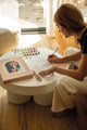 Woman painting the Parisian Door paint by numbers kit on a round white coffee table in a sunlit living room